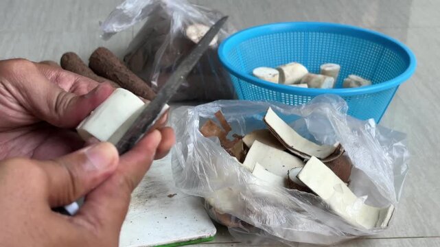 Hands are peeling and slicing cassava (Manihot esculenta) using a sharp knife. The peeled cassava pieces are placed in a blue basket to hold the slices, while the peels are collected in a plastic bag.