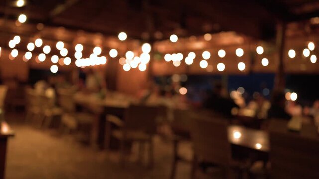 Blurred evening outdoor restaurant scene with warm hanging light bulbs and silhouettes of people dining in tropical resort.