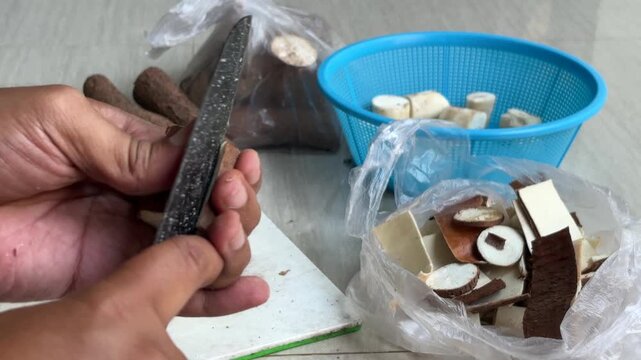 Hands are peeling and slicing cassava (Manihot esculenta) using a sharp knife. The peeled cassava pieces are placed in a blue basket to hold the slices, while the peels are collected in a plastic bag.
