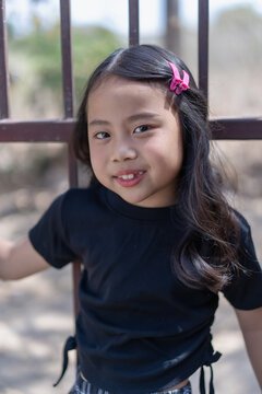 A close-up outdoor portrait capturing the smiling face of a young Asian girl with a missing front tooth, wearing a black top and dark patterned pants, accessorized with a bright pink hair clip