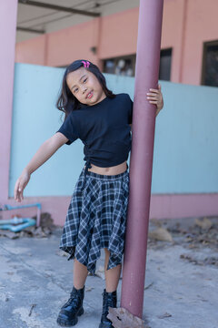 Cute little Asian girl wearing a black t-shirt and plaid skirt standing outdoors, holding a purple pole and looking at the camera with a sweet smile. Portrait of a happy child capturing a lovely momen
