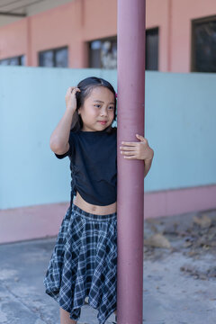 Cute little Asian girl wearing a black t-shirt and plaid skirt standing outdoors, holding a pole while touching her hair with a sweet smile. Portrait of a happy child capturing a lovely moment