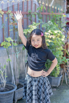 Happy young girl raising her hand outdoors in a garden, smiling at the camera in bright sunlight. Childhood, fashion, lifestyle, summer, nature, playful activity, confidence, and joyful youth concept