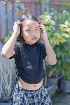 Young girl adjusting her hair outdoors in bright sunlight, wearing a black shirt and plaid skirt, with a natural portrait expression. Childhood, fashion, lifestyle, summer, and happy youth outdoors.