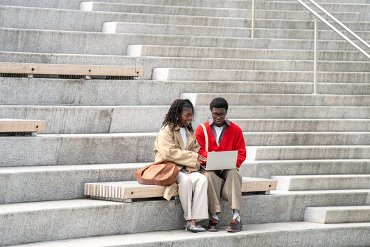African American friends use laptop on outdoor city steps, black couple students discussing coursework and sharing ideas while dressed in fashionable casual outfits during collaborative study session.