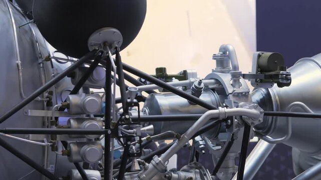 Close-up of vintage rocket engine on display. Metal pipes and black dome visible. Background shows blueprints and industrial equipment