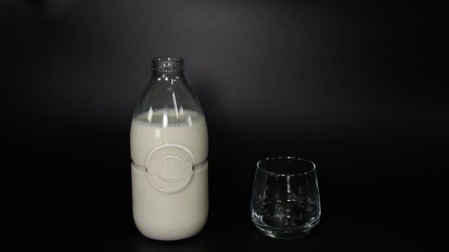 A person pouring fresh organic cow's milk from a transparent glass bottle into an empty glass on a black background.