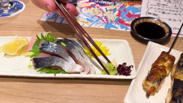 Person uses chopsticks to add wasabi to sashimi on a plate with grilled skewers nearby.