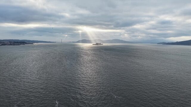 Aerial tracking view moving forward over open bay water toward Alcatraz Island, dramatically lit by breaking sun rays under a heavy overcast storm sky