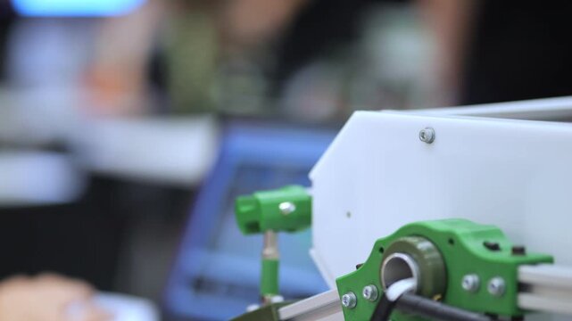 Child's hand operates small green robotic car with orange tire. Focused shot on mechanical parts. Background blurred, showing classroom setting