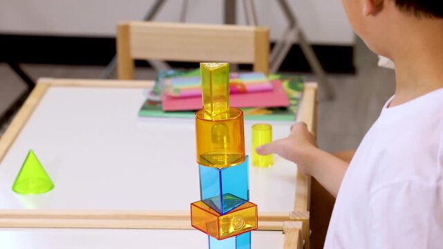 Young boy actively engages in learning geometry by carefully stacking colorful 3d shapes on white table during a bright day.