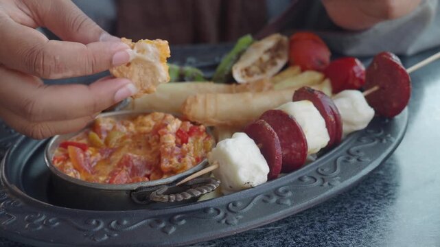 Hands dipping bread into turkish breakfast shakshuka with meat skewers and pastries on traditional platter in close up cinematic motion