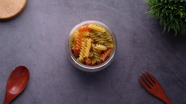 Hands dropping colorful fusilli pasta into glass bowl on dark tabletop in slow motion