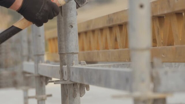 Close up detail shot showing builder hands wearing protective black gloves using heavy hammer securing metal scaffolding joints formwork upon construction development site