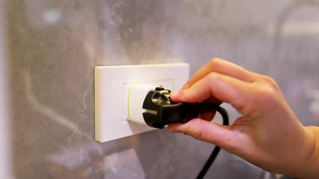 Close up hand plugging black electric power cord into universal travel adapter socket in modern home interior.