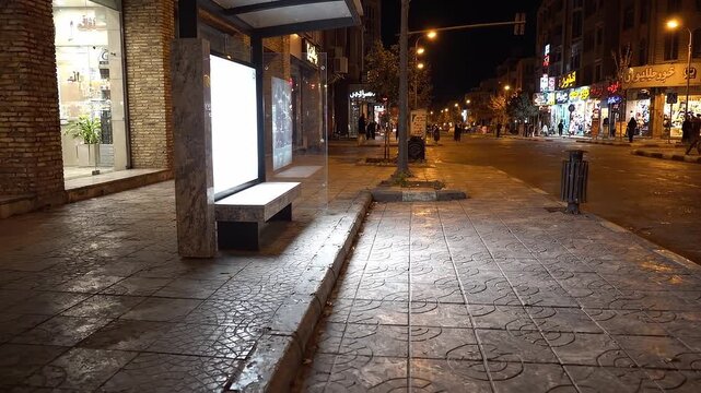Empty city street at night with illuminated bus stop and blank billboard. Urban sidewalk with glowing shop windows and street lamps.