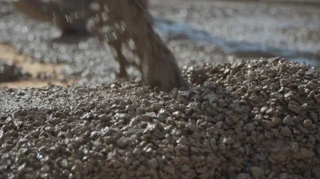 Close up slow motion shot capturing heavy wet concrete pouring onto gravel base while worker spreads cement mixture for new building foundation