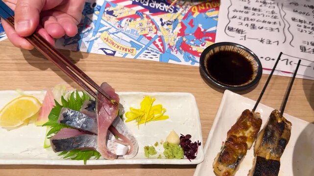 A close up shot of someone picking up a slice of fresh fish sashimi with chopsticks and dipping it in soy sauce in a japanese restaurant.
