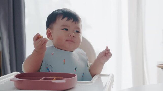 Asian baby boy eating boiled corn on high chair, BLW (baby-led weaning) concept, healthy solid food introducing to infant at home