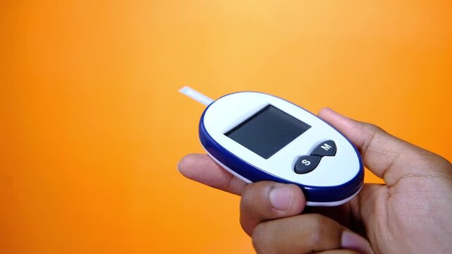 Person holding blood glucose meter with test strip inserted against orange background during medical checkup