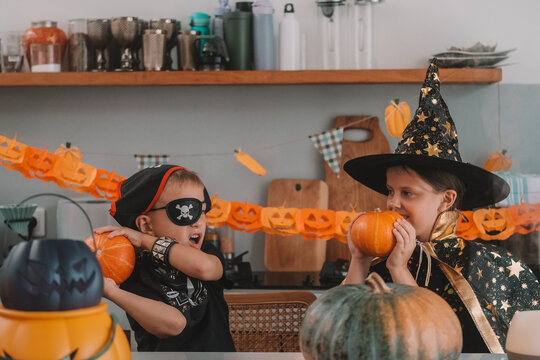 Portrait of a girl and her a little brother celebrating Halloween and decorating pumpkins in the kitchen. They look scary in witch and pirate costumes. Kids Halloween party.