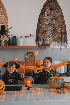 Portrait of a girl and her a little brother celebrating Halloween and decorating pumpkins in the kitchen. They look scary in witch and pirate costumes. Kids Halloween party.