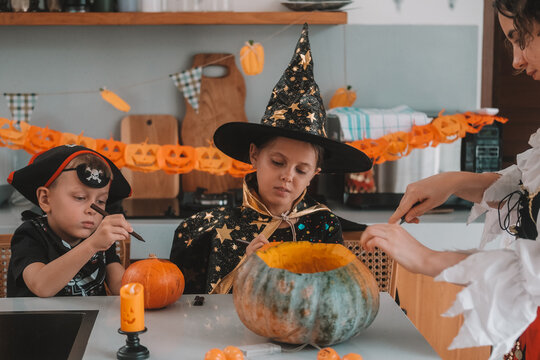 A portrait of a young girl and her younger brother celebrating Halloween and carving pumpkins in the kitchen with their mother's help. They are dressed in witch and pirate costumes. 