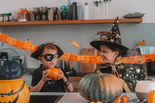 Portrait of a girl and her a little brother celebrating Halloween and decorating pumpkins in the kitchen. They look scary in witch and pirate costumes. Kids Halloween party.