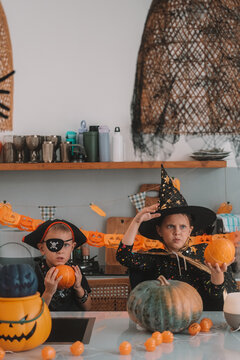 Portrait of a girl and her a little brother celebrating Halloween and decorating pumpkins in the kitchen. They look scary in witch and pirate costumes. Kids Halloween party.