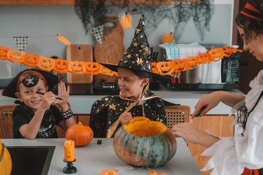 A portrait of a young girl and her younger brother celebrating Halloween and carving pumpkins in the kitchen with their mother's help. They are dressed in witch and pirate costumes. 