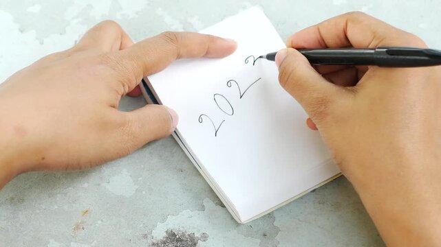 Person writing year 2023 on white notebook using black pen with hand motion