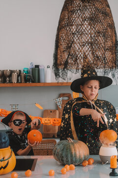 Portrait of a girl and her a little brother celebrating Halloween and decorating pumpkins in the kitchen. They look scary in witch and pirate costumes. Kids Halloween party.