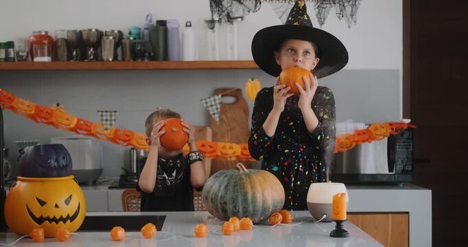 Portrait of a girl and her a little brother celebrating Halloween and decorating pumpkins in the kitchen. They look scary in witch and pirate costumes. Kids Halloween party.