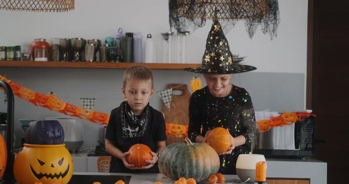 Portrait of a girl and her a little brother celebrating Halloween and decorating pumpkins in the kitchen. They look scary in witch and pirate costumes. Kids Halloween party.