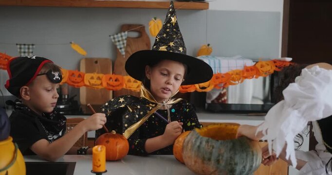 A portrait of a young girl and her younger brother celebrating Halloween and carving pumpkins in the kitchen with their mother's help. They are dressed in witch and pirate costumes. 