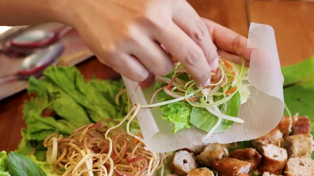 Close up of hands making Vietnamese spring rolls with fresh lettuce herbs and grilled meat on rice paper.
