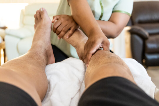 Close-up perspective view of masseuse performing traditional relaxing and therapeutic Thai foot and leg massage, pressing on all essential acupoints in nice ambient environment.