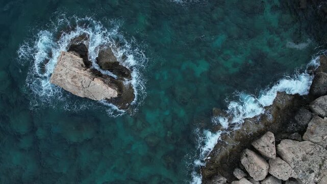 Drone aerial seascape of Azure sea waves breaking on a rocky shore and a solitary cliff, creating white foam