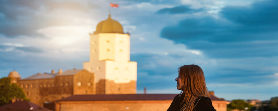 A young woman thoughtfully looks out at the Vyborg Castle during the golden hour as dramatic clouds roll in, suggesting an impending storm.