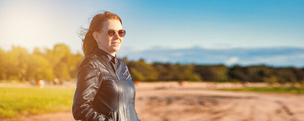 An adult woman stands confidently on the beach, wearing a leather jacket and sunglasses. The sunny day sets a cheerful mood, with a vibrant outdoor setting in the background. © Aleksandr