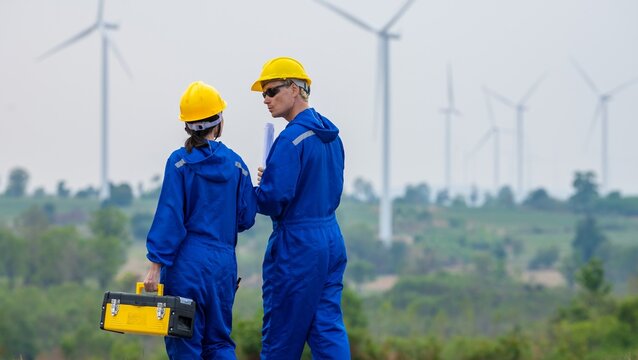 Team technician discussing wind turbine power generator sustainability performance inspection together at Renewable Energy Plant Male and female workers in coverall hold toolbox and walking site