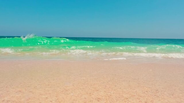 Slow motion low angle view of white sea foam washing ashore on golden sand beach under turquoise ocean and clear blue sky.
