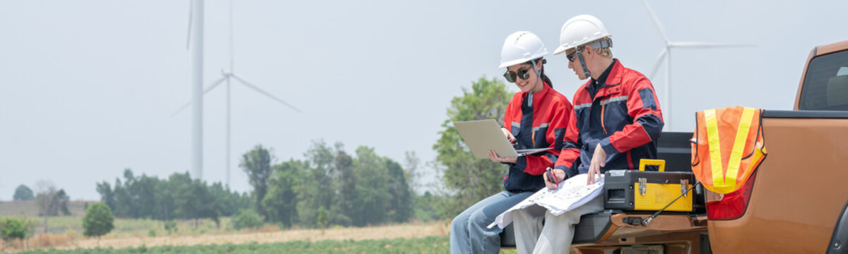 Engineers team sitting on car trunk tailgate SUV using laptop and blueprint renewable energy near wind turbine farm male and female technicians wear hardhats working on SUV at wind power plant