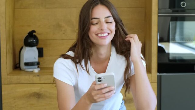Beautiful young woman with a radiant smile laughing sincerely while looking at her mobile phone, enjoying a funny video or message in her modern kitchen, showing genuine happiness