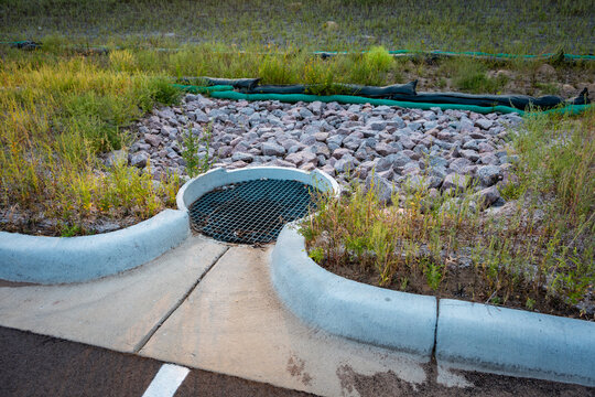 A modern precast concrete catch basin with an iron drainage grate, located at the edge of an asphalt parking lot with surrounding riprap rock