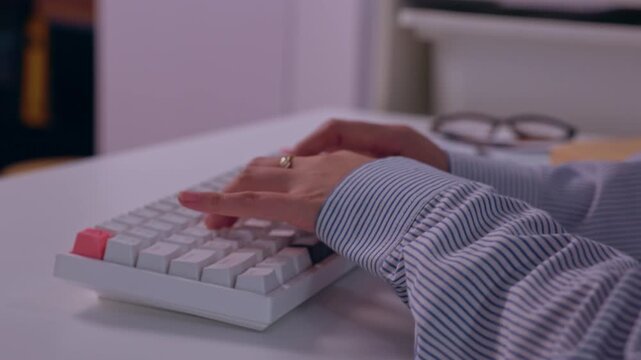Person typing white mechanical keyboard desk, striped shirt sleeve and wedding ring visible, focused computer work office, hands creating productive work scene, eyeglasses softly blurred background