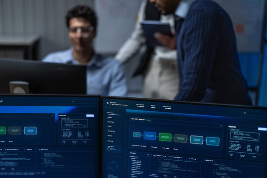 Desktop monitors in the foreground of a dark office sit lit on a shared desk with a coffee tumbler beside them in a blue lit engineering room, shot in low light.