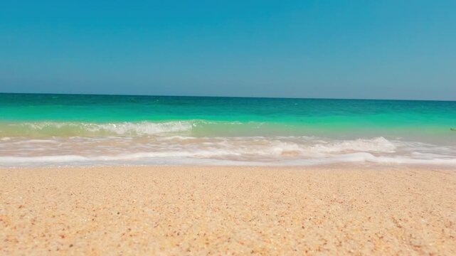 Slow motion low angle view of white sea foam washing ashore on golden sand beach under turquoise ocean and clear blue sky.

