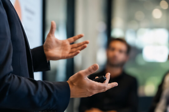 a speaker giving a presentation in a professional business setting, using a clicker and expressive hand gestures while an audience listens in the background.  seminar or leadership design element