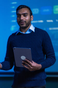 A South Asian software lead in dark knitwear holding a slim digital tablet tilts his head slightly while considering the figure on the screen in a professional corporate environment